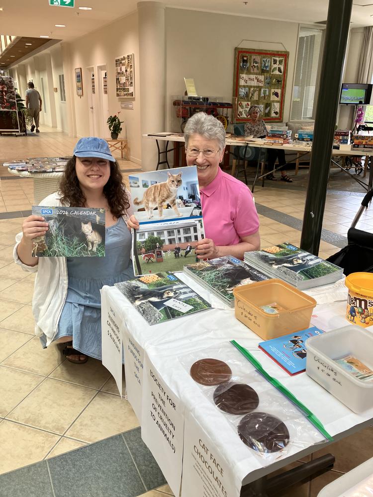 a young woman in a blue dress and cap and older woman in a bright pink top hold up copies of the SPCA’s 2026 calendar